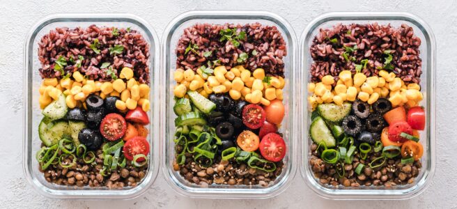 Three identical glass food storage containers holding prepared wild rice, sweetcorn, black olives, tomatoes, cucumber, spring onion, green lentils and herbs.
