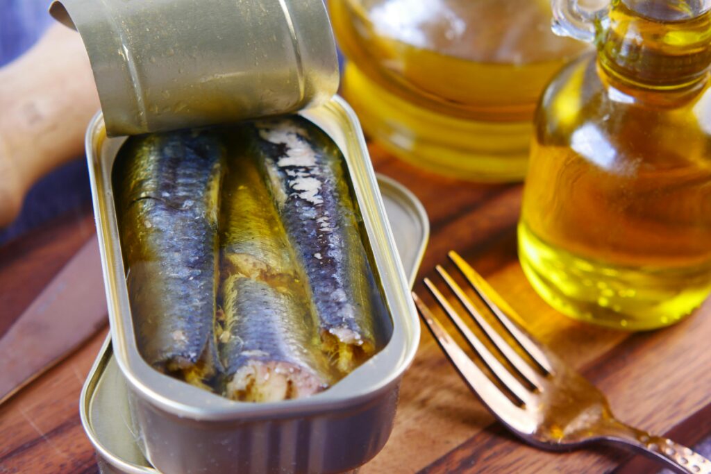 Open can of sardines in olive oil, beside glass jars of olive oil.
